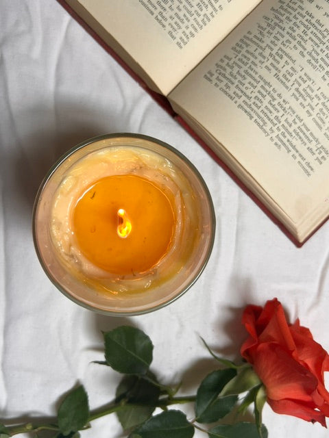 Candle in a glass holder with an open book and a red rose on a white surface