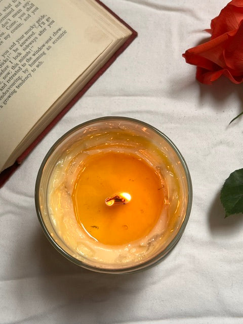 Candle in a glass jar with a lit wick, placed on a white fabric surface next to a book and red flower.